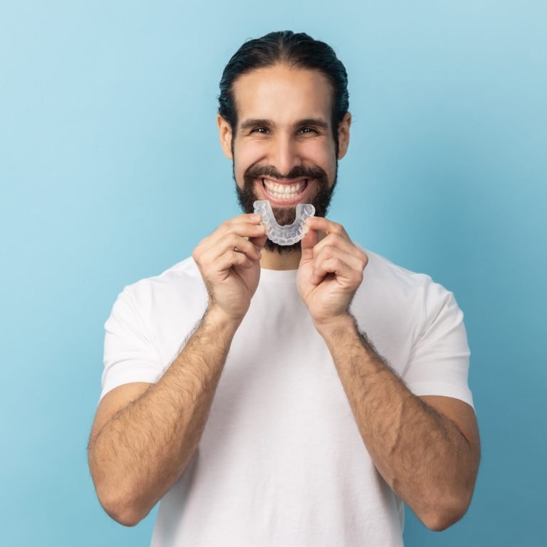 Portrait of man with beard in white T-shirt using teeth whitening braces, dental aligner retainer, dental clinic for beautiful teeth, treatment course. Indoor studio shot isolated on blue background.