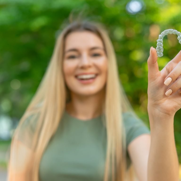 Happy girl in green t-shirt holding removable braces or aligners for straightening and whitening teeth on green background
